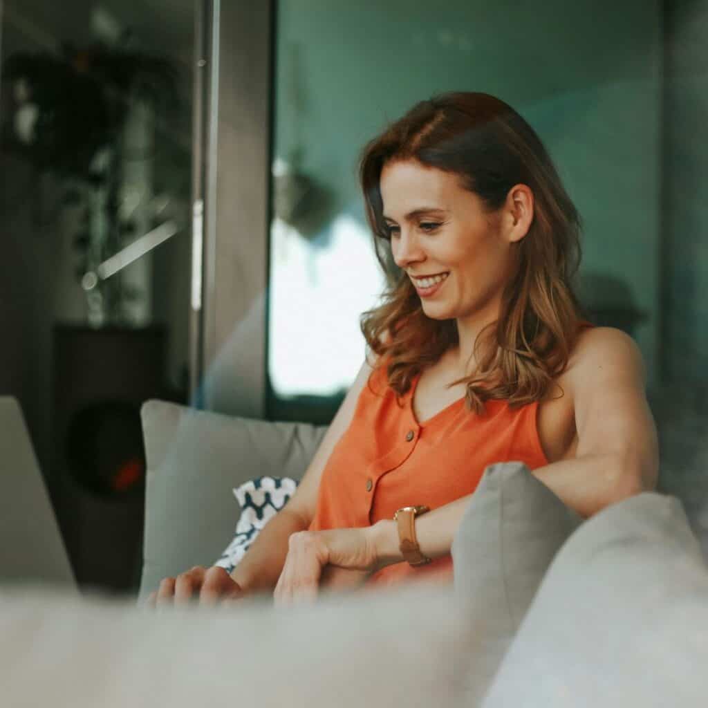 A smiling woman with reddish-brown hair, wearing an orange top, sits on a gray sofa, looking down at a device.