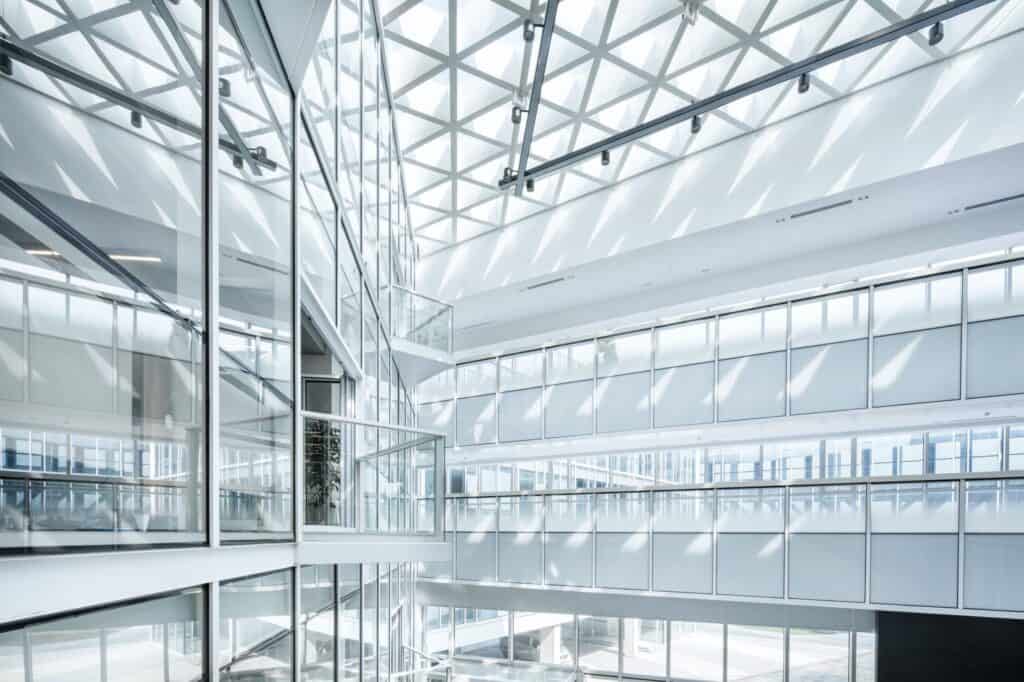 A bright, multi-story modern atrium with extensive glass walls, glass railings, and a geometric grid skylight ceiling casting triangular shadows.