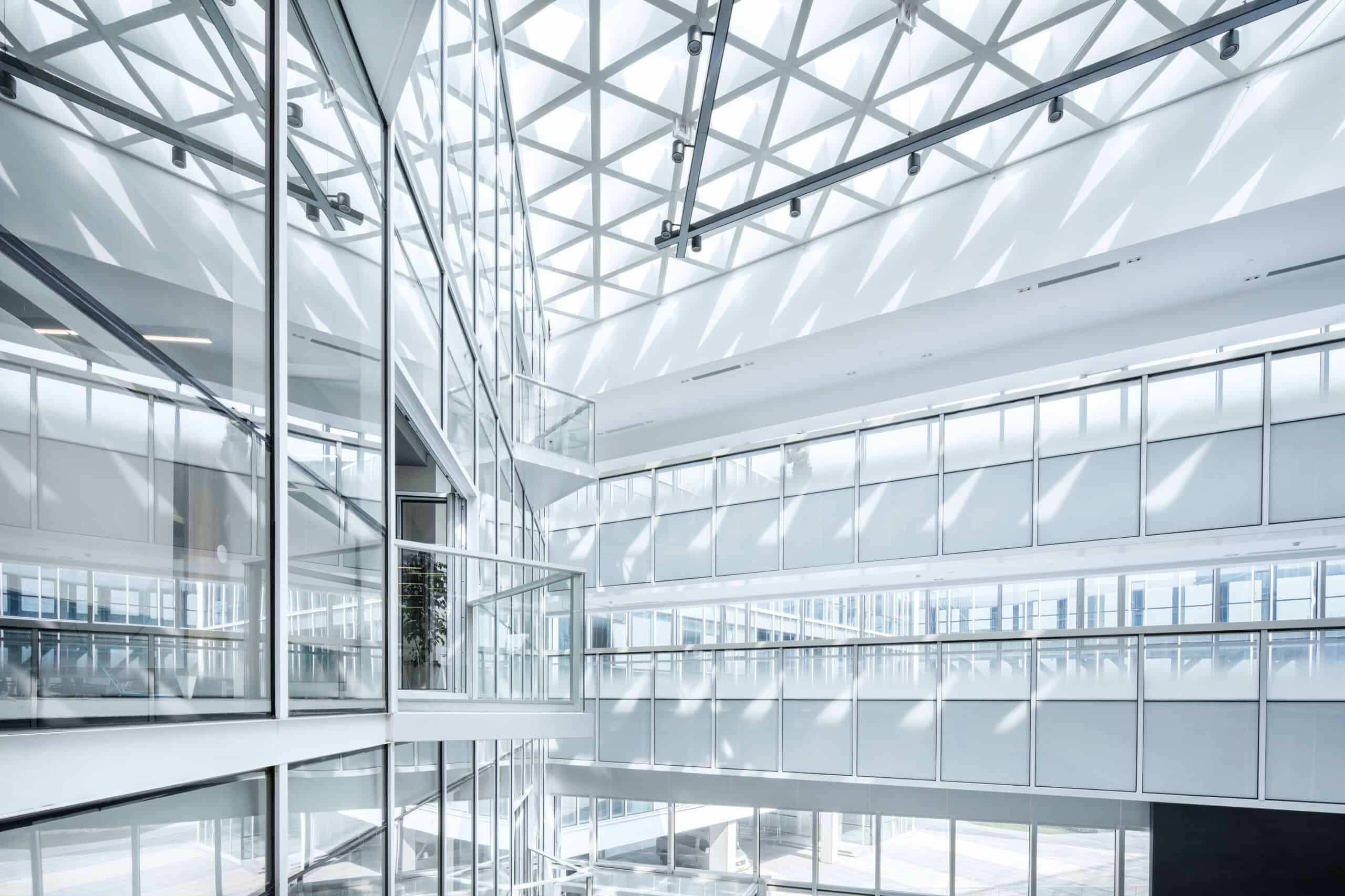 A bright, multi-story modern atrium with extensive glass walls, glass railings, and a geometric grid skylight ceiling casting triangular shadows.