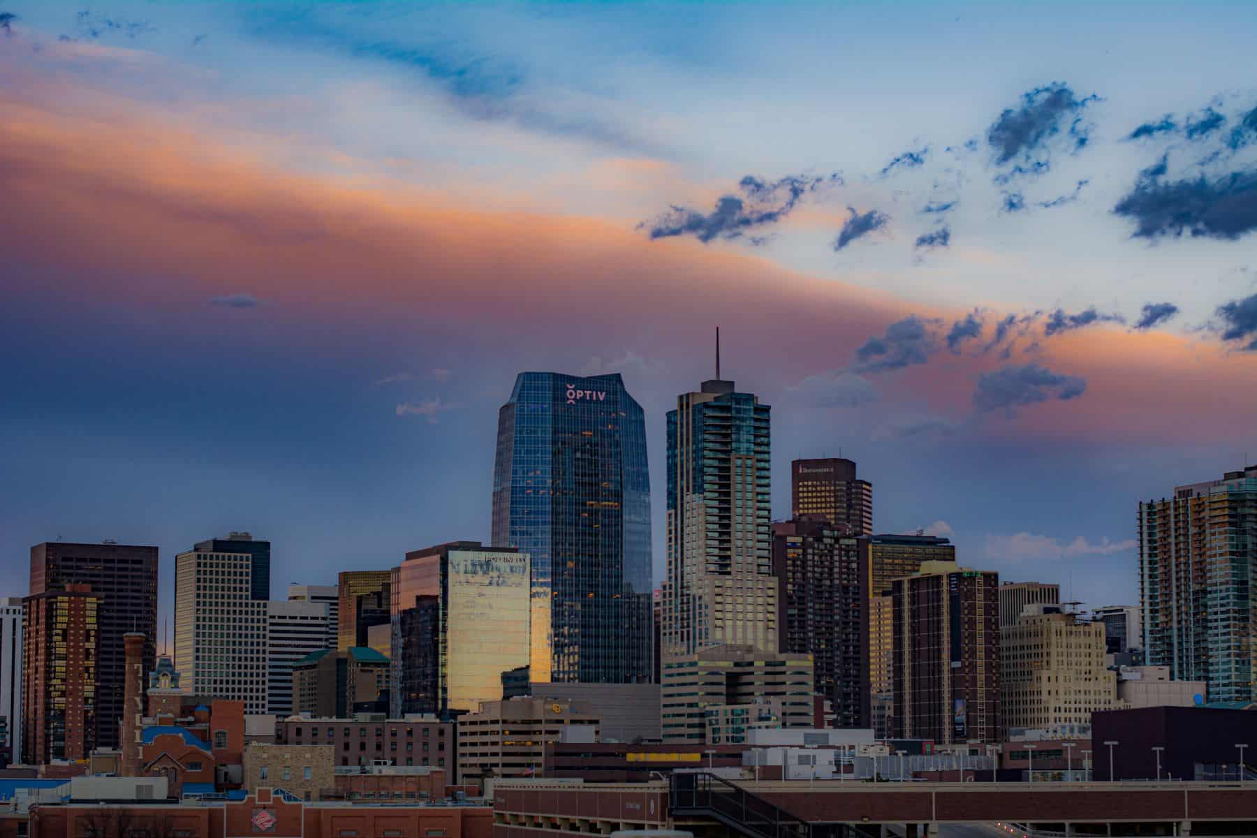 A cityscape at dusk with a vibrant orange, pink, and blue sky over numerous buildings, including a prominent skyscraper with an 'OPTIV' logo.