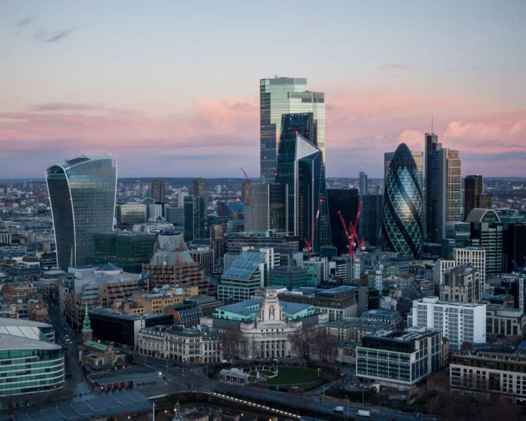 An aerial view of the London skyline at dusk, featuring the Walkie-Talkie and Gherkin skyscrapers against a pink and purple sky.