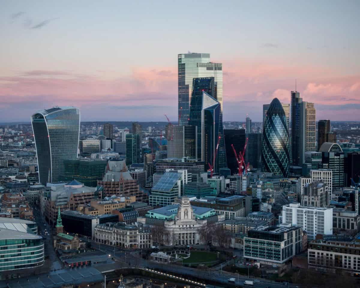An aerial view of the London skyline at dusk, featuring the Walkie-Talkie and Gherkin skyscrapers against a pink and purple sky.