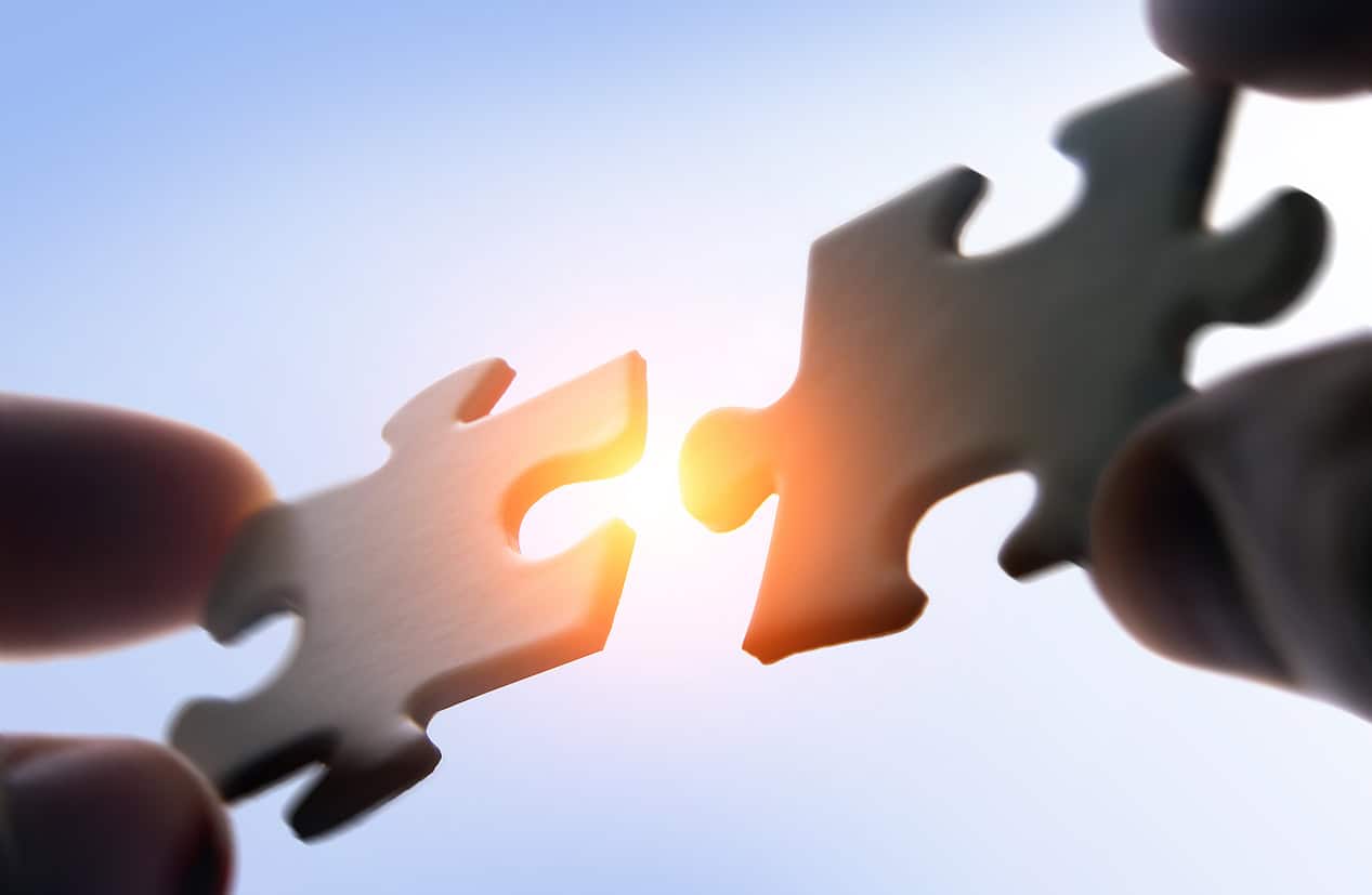 Two light brown puzzle pieces held by fingers, about to connect, with bright orange light shining between them against a blue sky background.