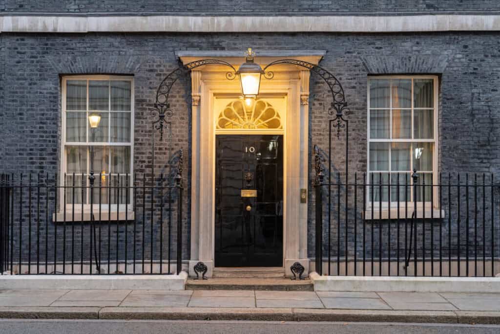 The black door of 10 Downing Street with the number 10, illuminated by a lantern, flanked by two windows and a black iron fence.