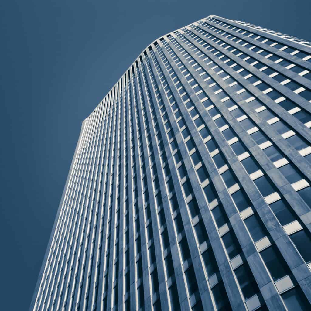 Low-angle view of a tall, modern building facade with repetitive dark windows and light panels, set against a clear blue sky. The image is monochromatic blue.