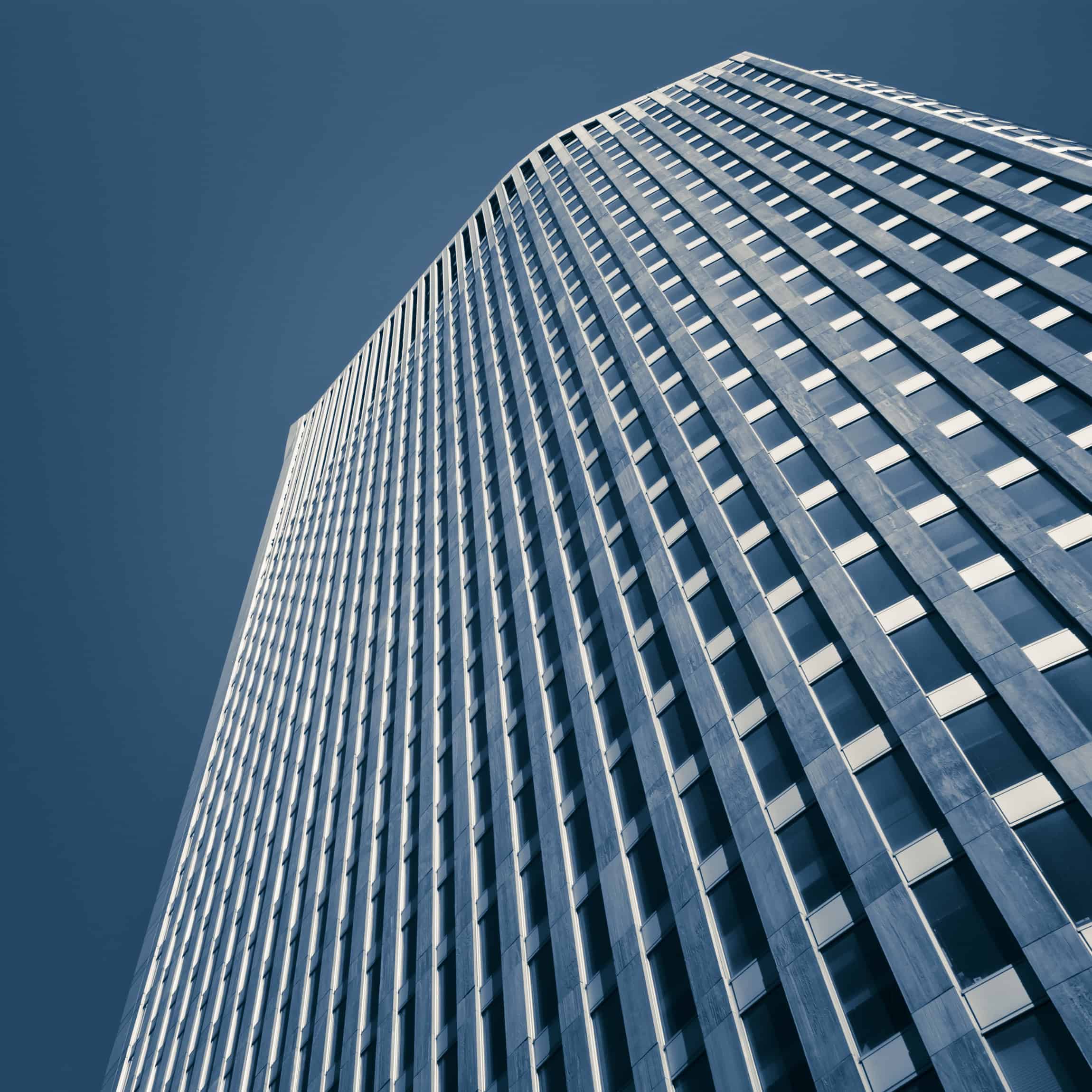 Low-angle view of a tall, modern building facade with repetitive dark windows and light panels, set against a clear blue sky. The image is monochromatic blue.