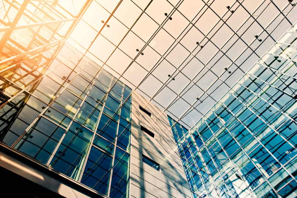 Upward view of a modern building's interior, featuring a vast glass ceiling and walls with a metal grid, illuminated by bright sunlight.