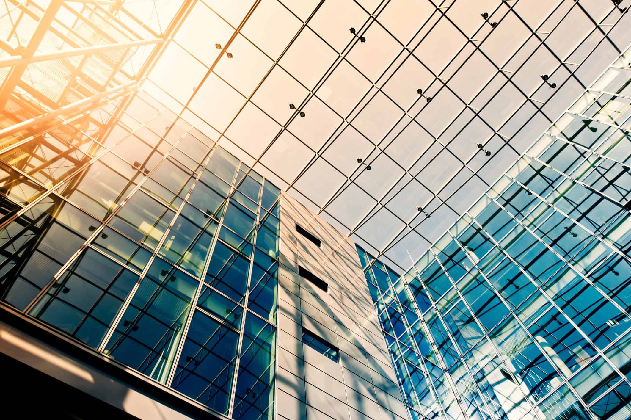 Upward view of a modern building's interior, featuring a vast glass ceiling and walls with a metal grid, illuminated by bright sunlight.