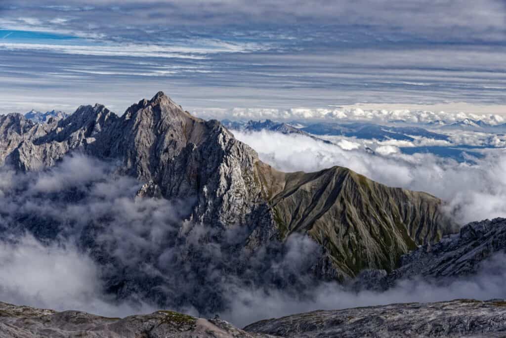 Panoramic view of rugged mountain peaks surrounded by a thick layer of white clouds, with layered grey and white clouds in the sky above.