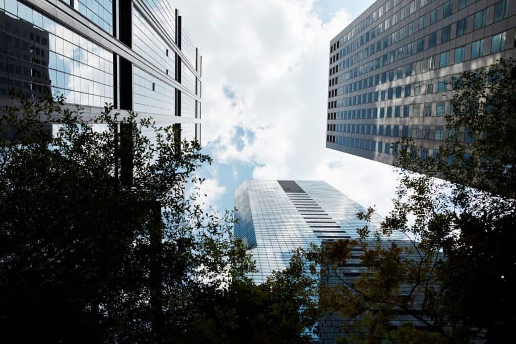 Low-angle view of three modern skyscrapers with glass and grey facades, framed by dark green tree leaves, against a cloudy sky.