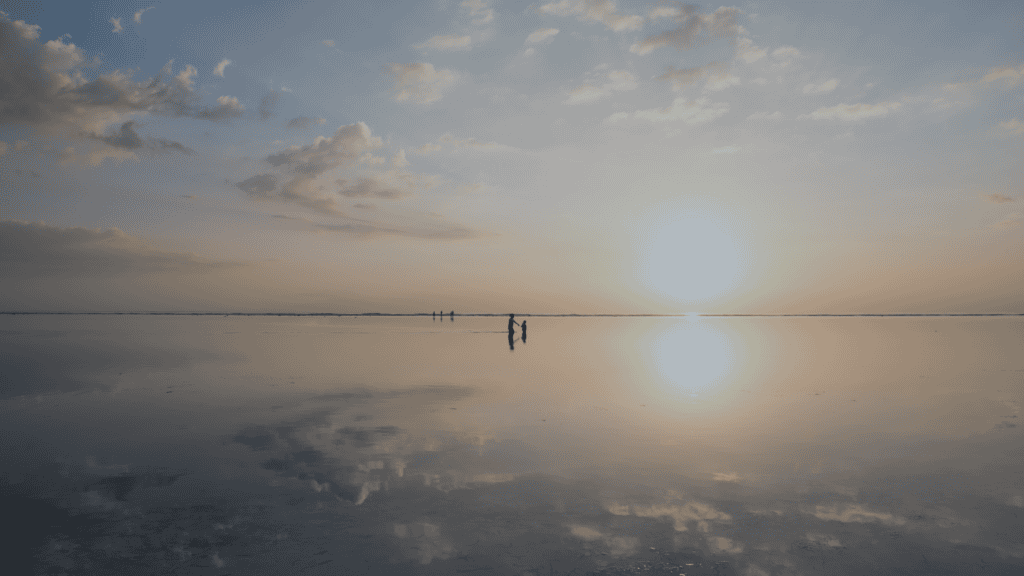 A wide shot of a sunset over calm, shallow water reflecting the sky and sun. Silhouetted figures stand in the distance.