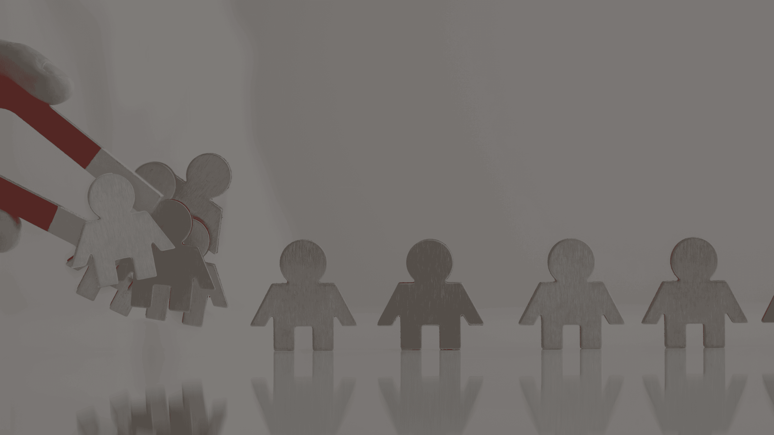 A hand holds a red and white magnet attracting several metallic human figures, while a line of five similar figures stands on a reflective surface.