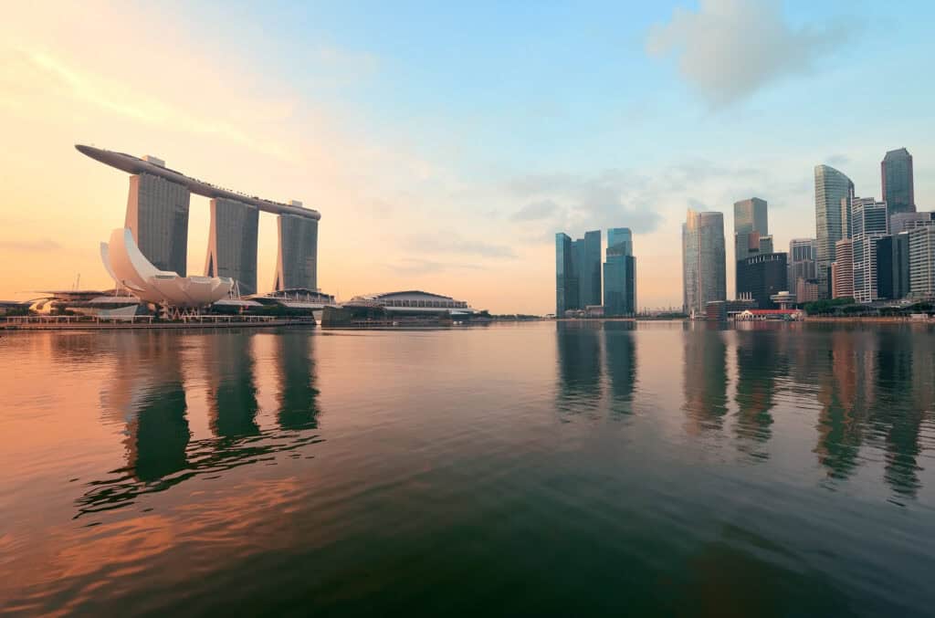 Panoramic view of Singapore's waterfront cityscape at sunrise, showing Marina Bay Sands and skyscrapers reflected in the bay.