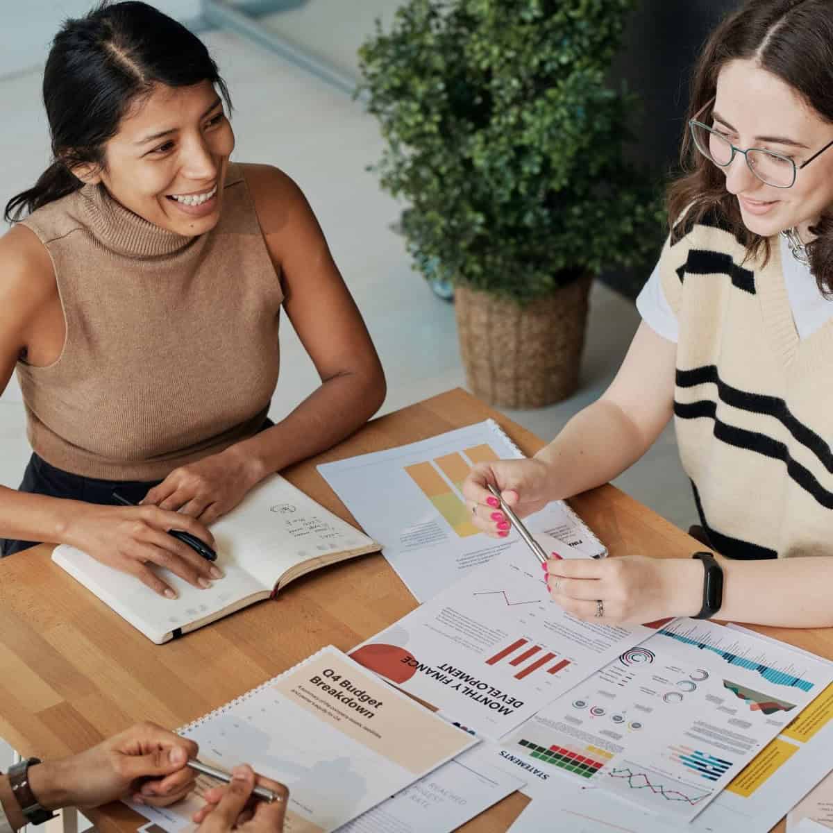 Three people at a wooden table reviewing business documents with charts and graphs. One woman smiles, another points at papers with a pen.