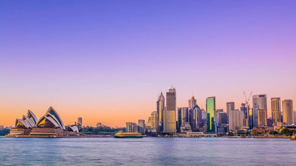Panoramic view of Sydney skyline at sunset, showing the Opera House and skyscrapers reflecting a purple and orange sky over calm water.