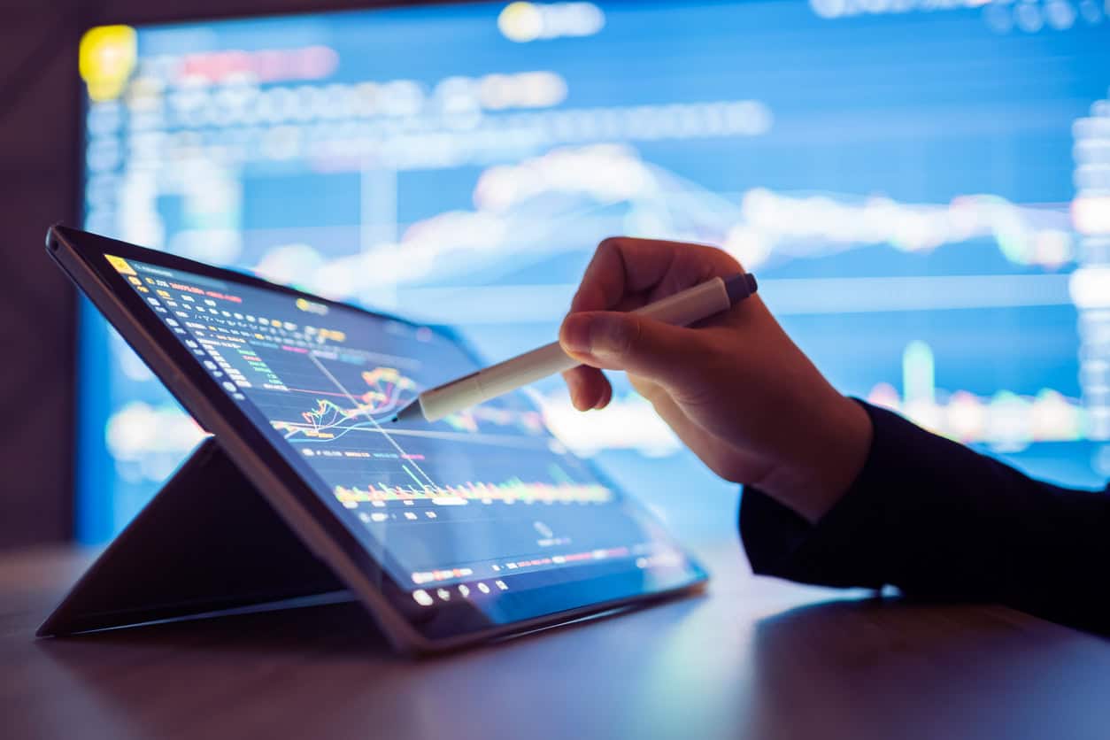 A hand holding a stylus points at financial charts on a tablet screen, with another screen showing charts in the background.