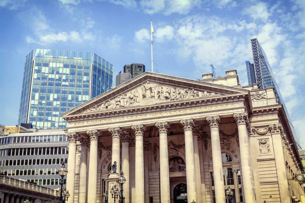 The classical facade of the Royal Exchange building with ornate columns and sculptures, flanked by modern glass skyscrapers under a blue sky.
