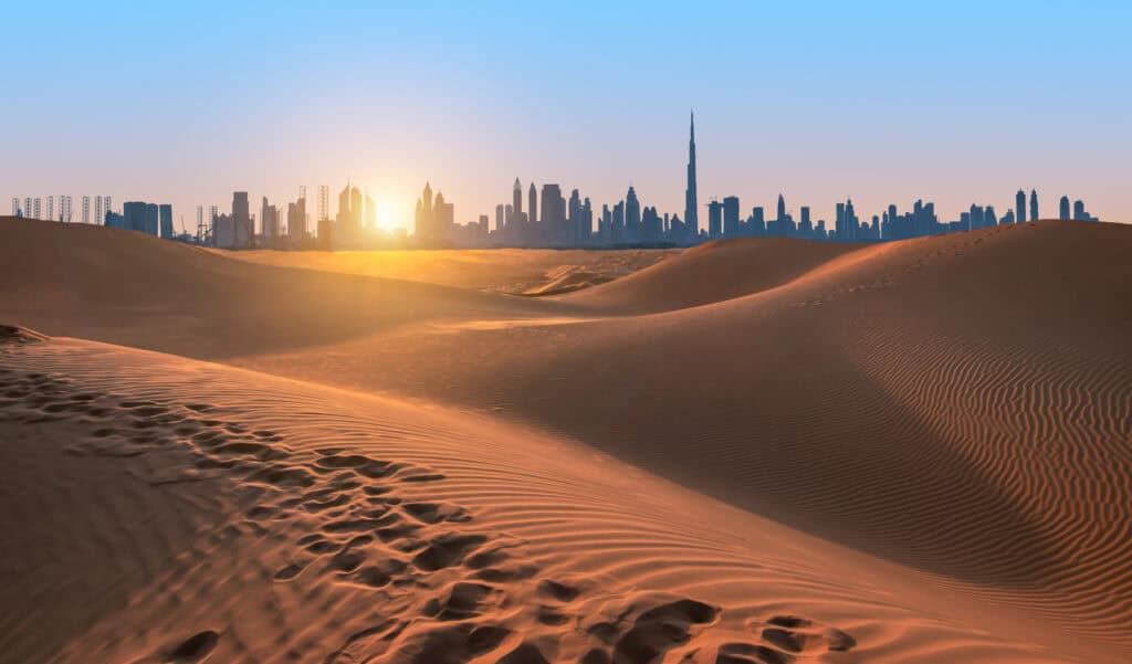 A desert landscape with rolling sand dunes in the foreground, showing footprints. In the background, a modern city skyline is silhouetted against a vibrant orange and blue sky, with the sun low on the horizon.
