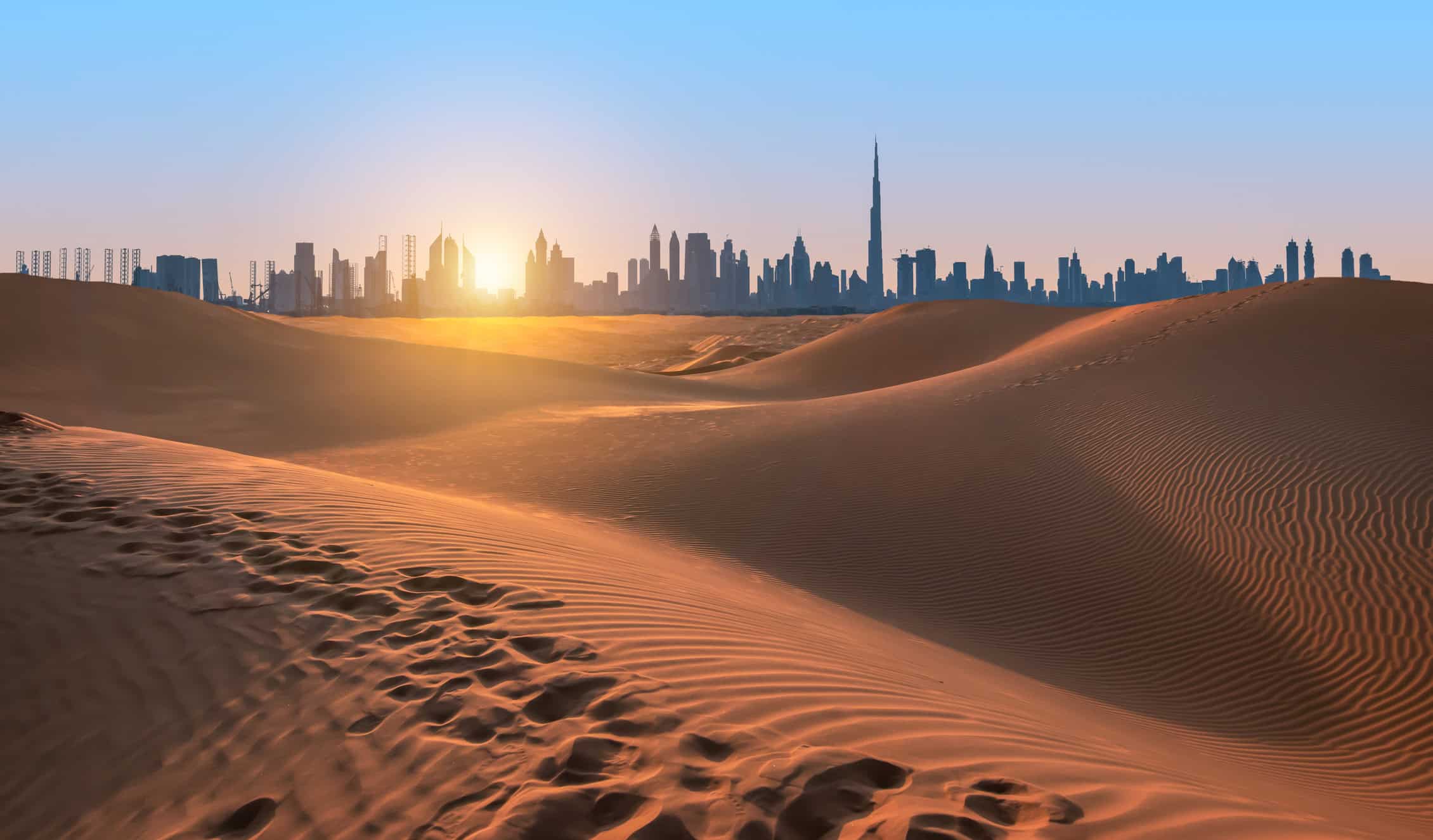 A desert landscape with rolling sand dunes in the foreground, showing footprints. In the background, a modern city skyline is silhouetted against a vibrant orange and blue sky, with the sun low on the horizon.