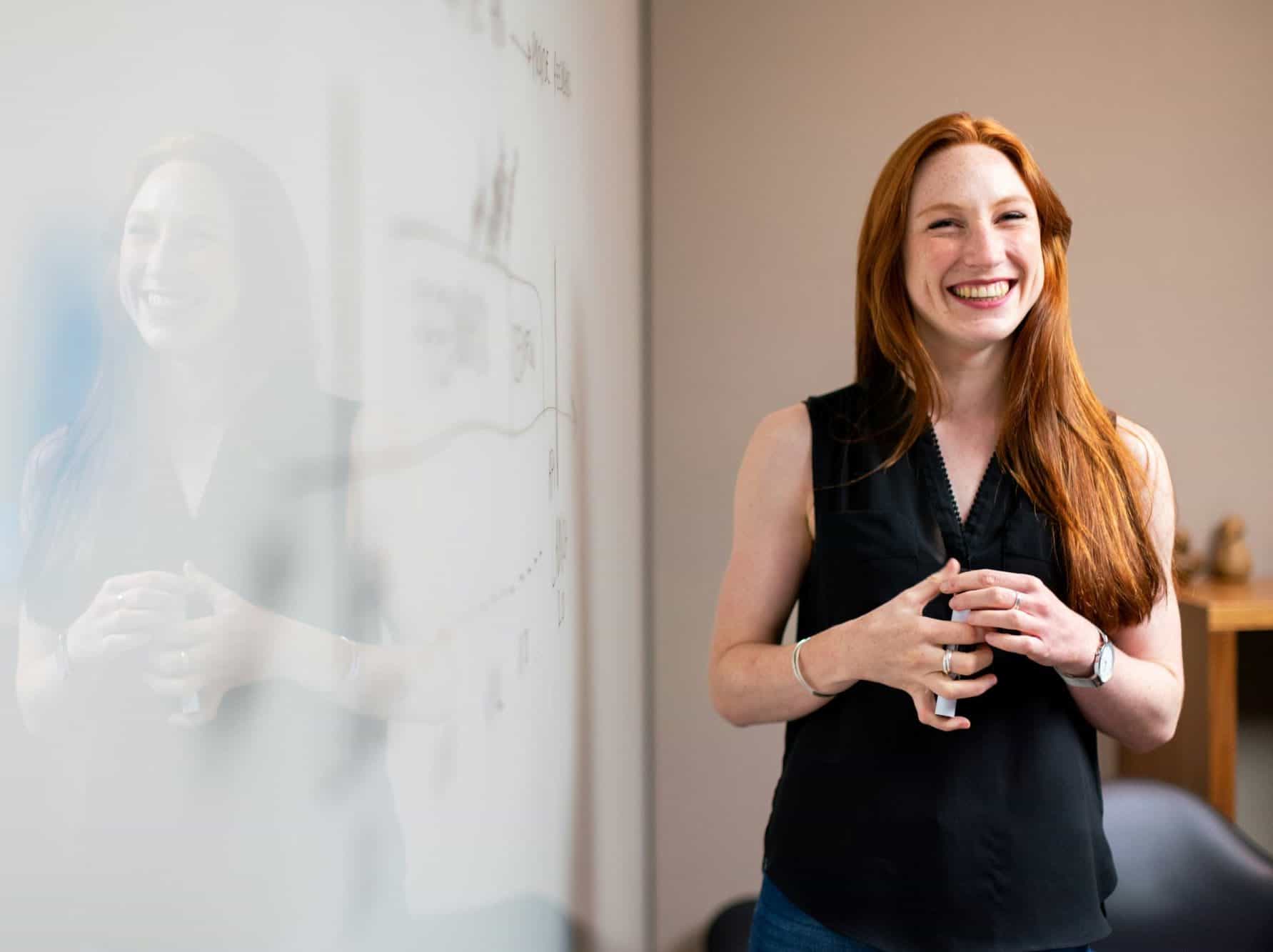 A red-haired woman in a black sleeveless top smiles broadly while standing next to a whiteboard with diagrams.