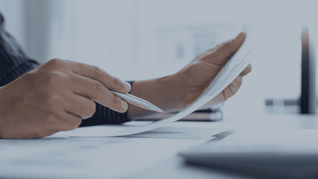 Close-up of hands holding a pen and a document with blue bar charts on a desk with other papers and a laptop.