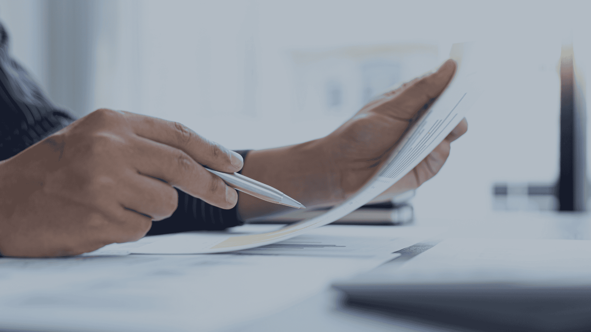 Close-up of hands holding a pen and a document with blue bar charts on a desk with other papers and a laptop.