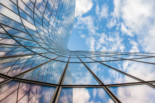 Low-angle view of a curved glass skyscraper reflecting a blue sky with white clouds.