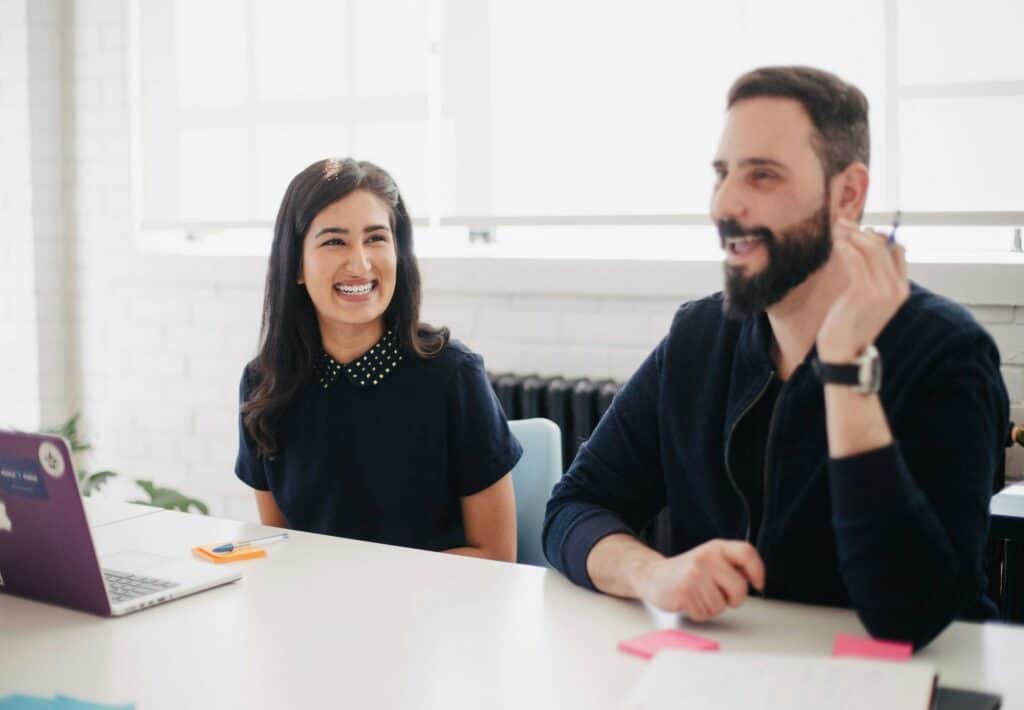 Two people, a woman and a man, smiling at a table with a laptop and notes.