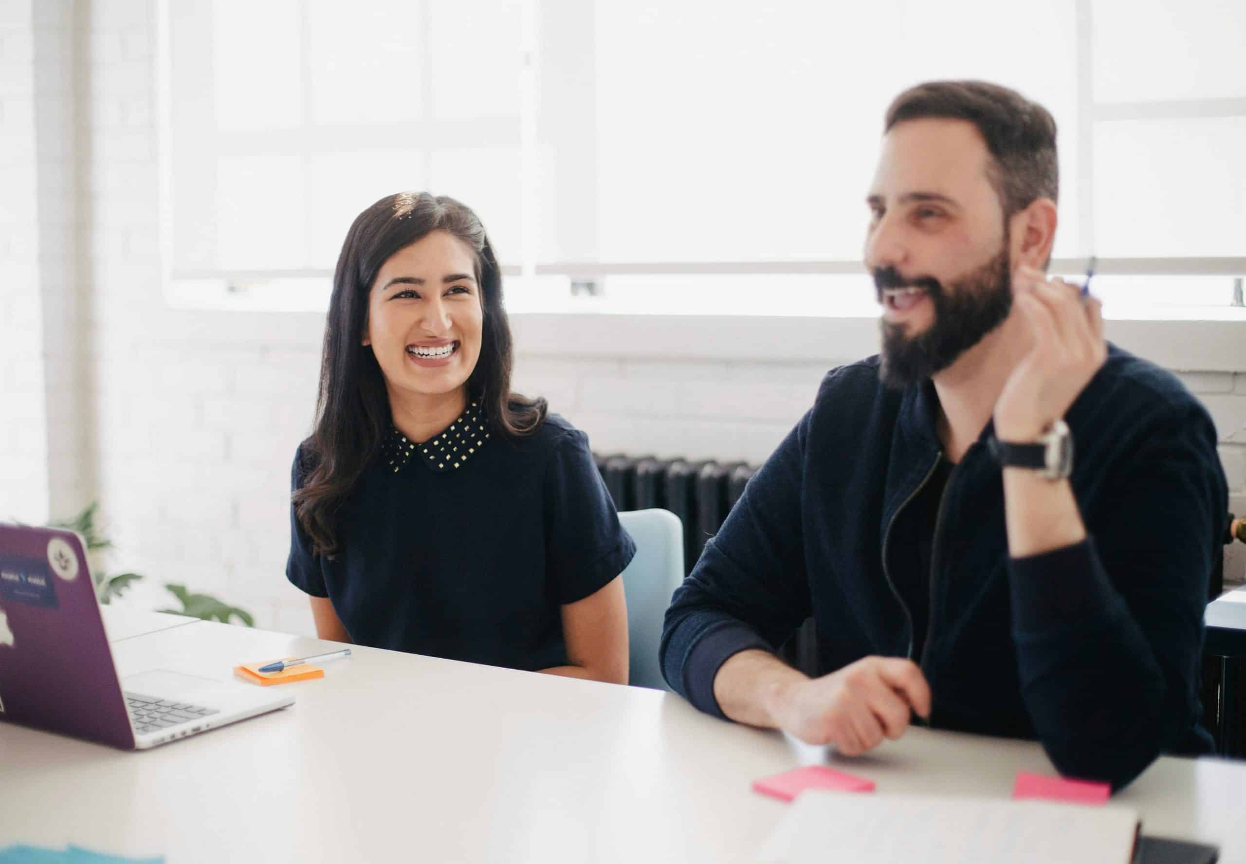 Two people, a woman and a man, smiling at a table with a laptop and notes.