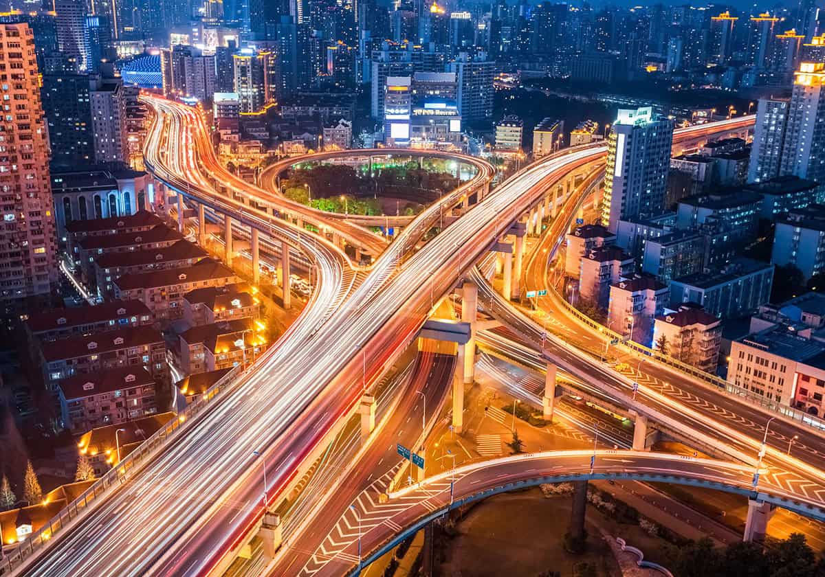 Aerial long-exposure view of a multi-level highway interchange at night, with bright red and white light trails from moving vehicles and illuminated city buildings in the background.