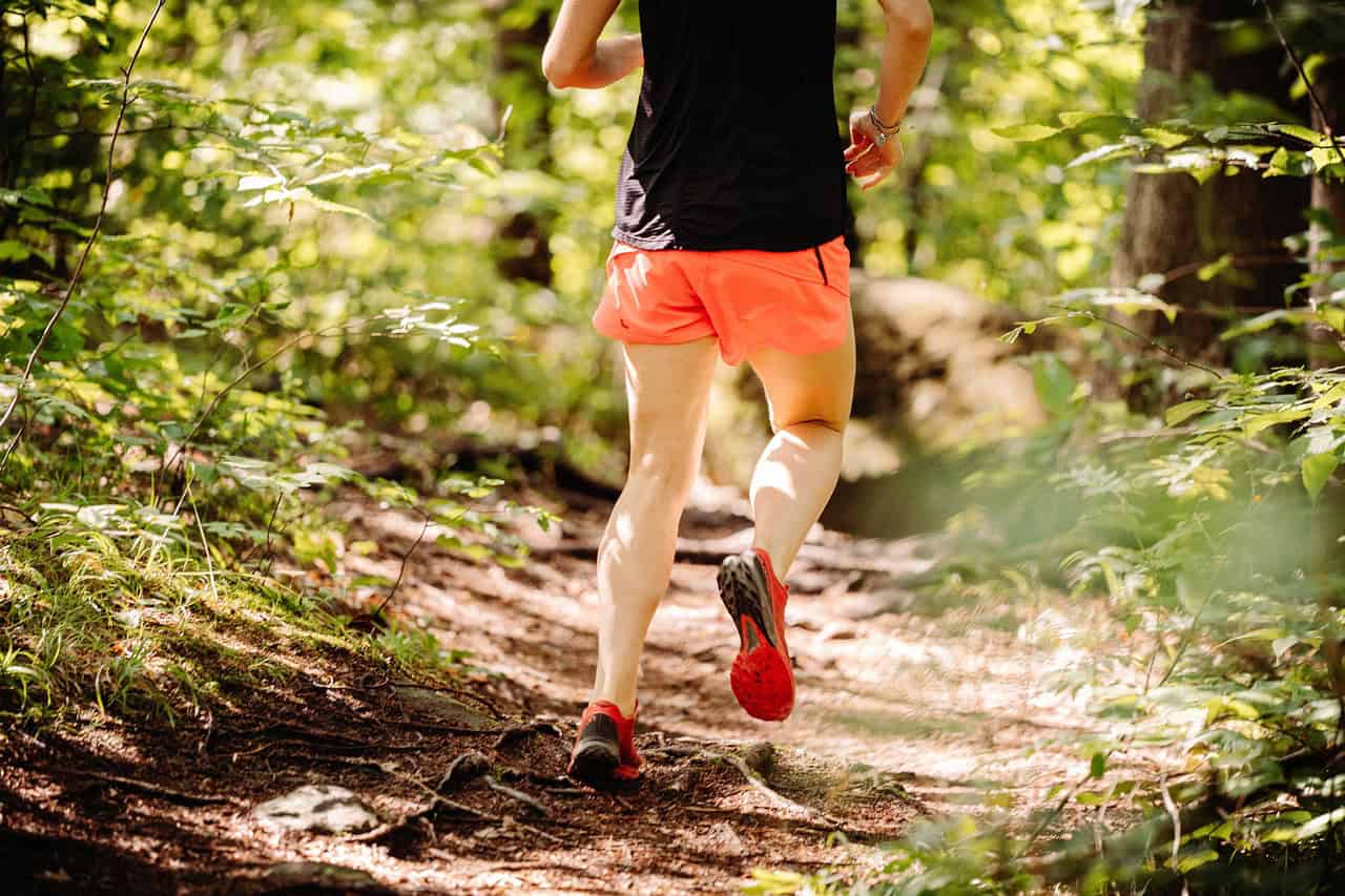 A person in orange shorts and a black top runs on a dirt trail in a forest, viewed from behind.