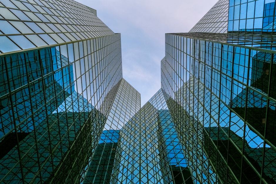 A low-angle view of multiple glass skyscrapers, showing their reflective blue and teal facades and grid patterns against a light sky.
