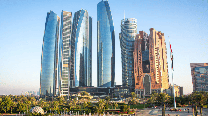 A panoramic view of modern skyscrapers, including glass towers and a traditional-style building, surrounded by palm trees and a fountain, with the UAE flag flying.
