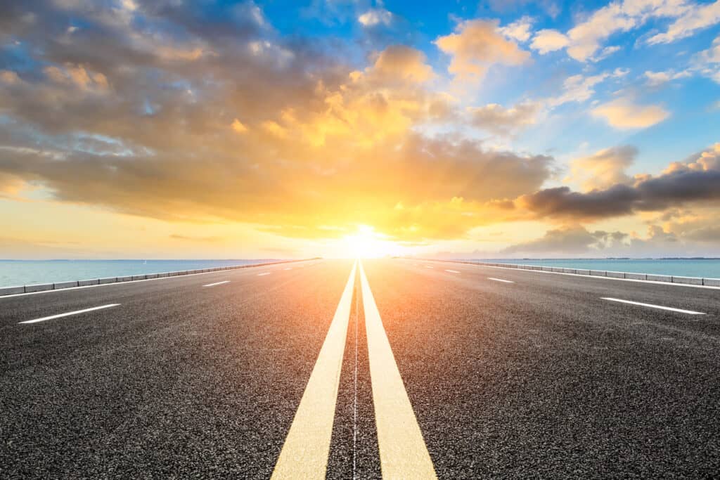 A straight asphalt road with double yellow lines extends towards a bright sunset over the ocean, flanked by water on both sides.