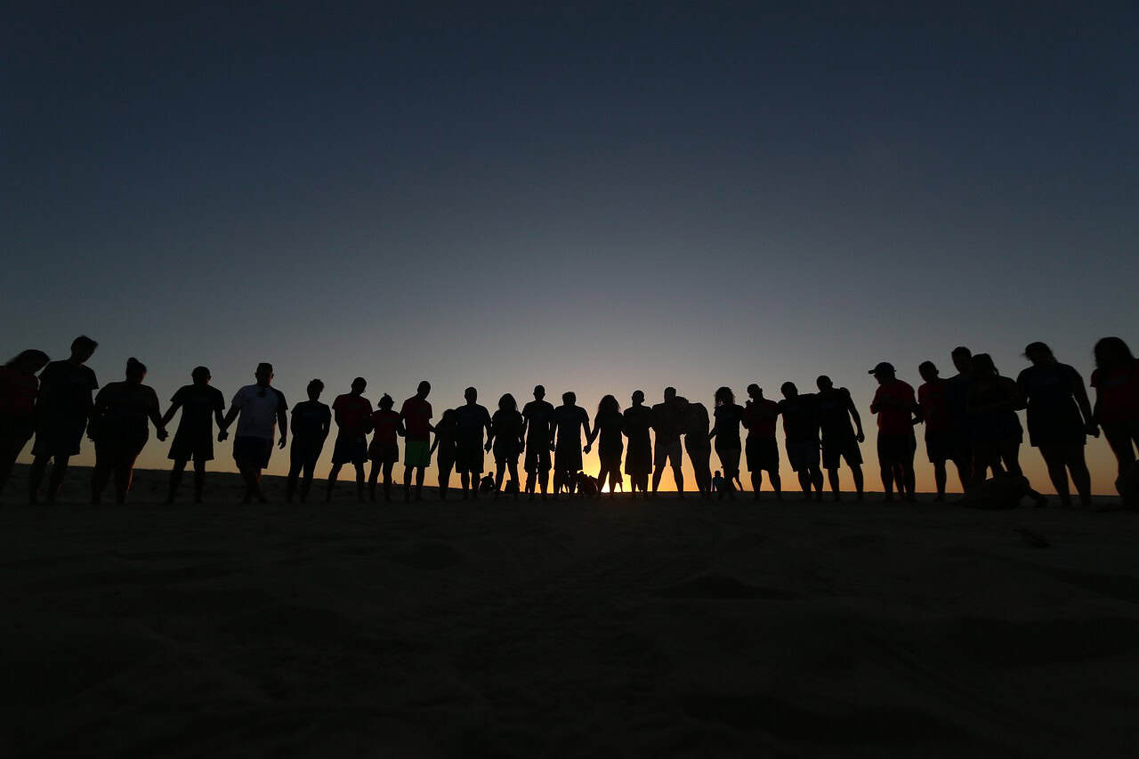 A large group of people silhouetted against a bright orange sunset on a sandy dune.