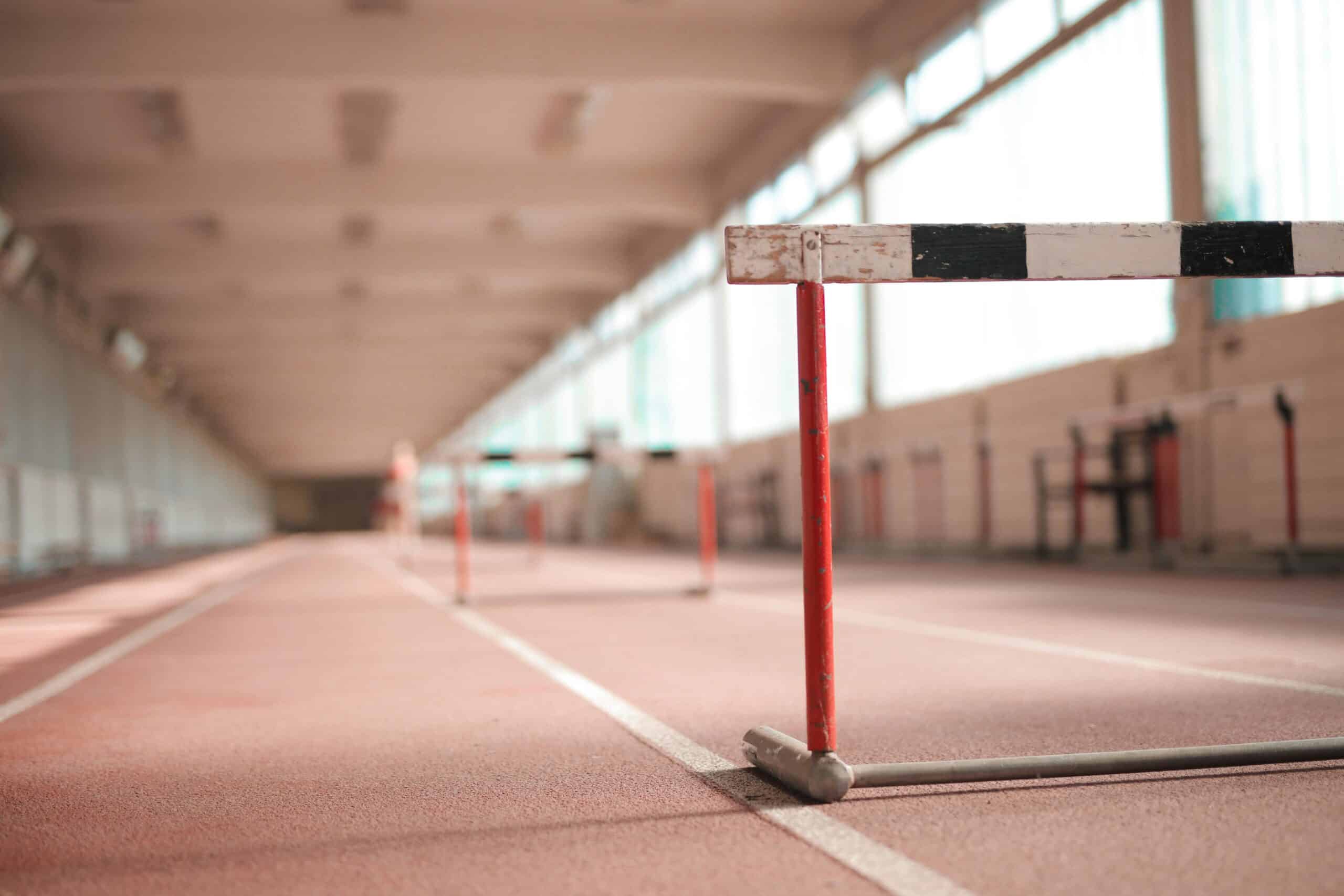 A close-up of a red and white hurdle on a reddish-brown indoor running track, with more hurdles blurred in the distance.