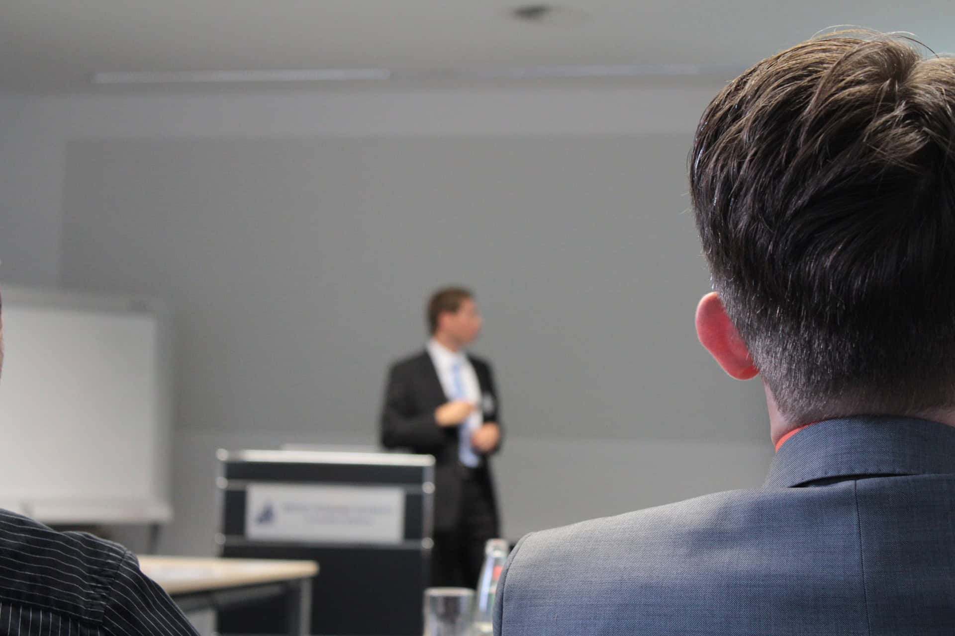 The back of a person's head and shoulder in a grey suit, facing a blurred presenter at a podium in a conference room.