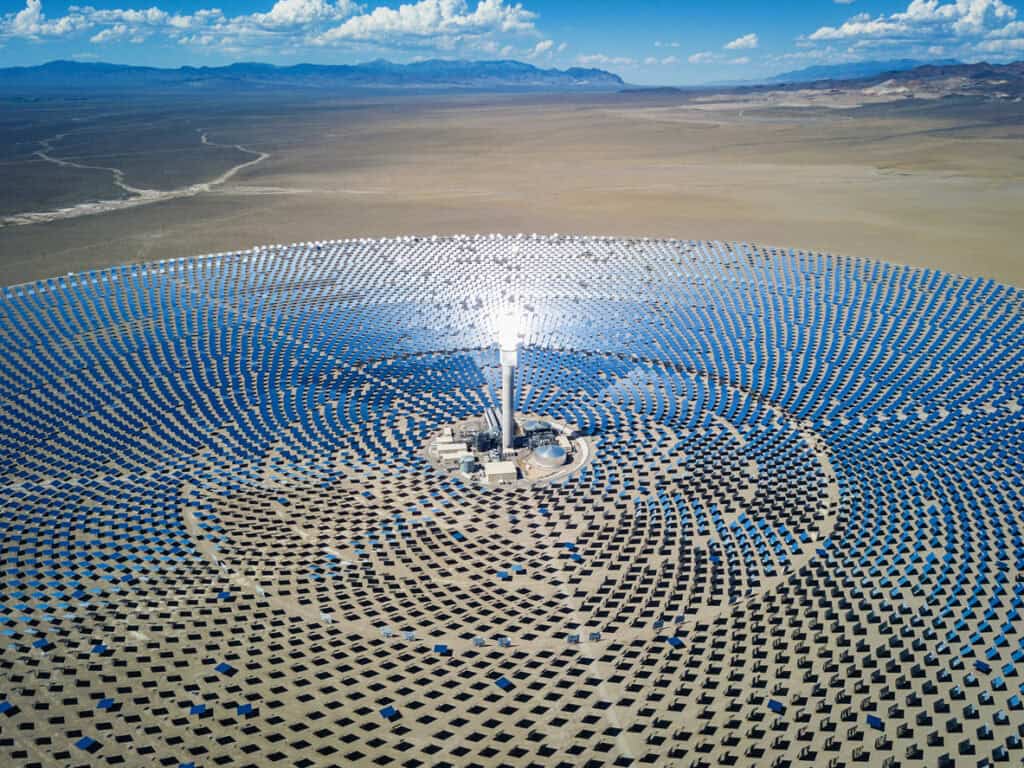 Aerial view of a large solar thermal power plant in a desert, featuring a central tower glowing brightly from concentrated sunlight reflected by thousands of surrounding mirrors.