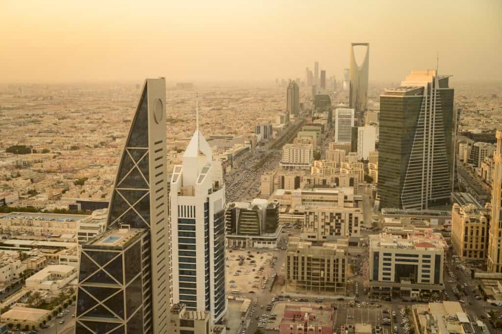 Aerial view of Riyadh cityscape at sunset, featuring prominent skyscrapers, a busy highway, and sprawling urban areas under a hazy, golden sky.