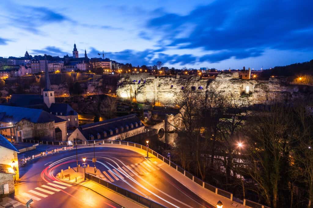 A long-exposure night photo of a city built on cliffs. A winding road with red and white light trails from vehicles is in the foreground. Illuminated cliffs rise in the midground, topped by a city with a church spire and other buildings. The sky is dark blue with clouds.
