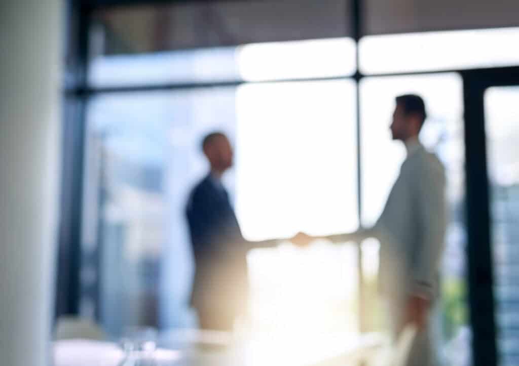 Blurred image of two men in suits shaking hands in front of a bright, sunlit window.