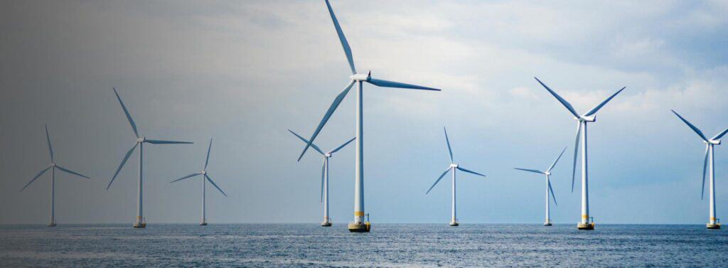 Ten large white offshore wind turbines with three blades each, standing in a blue ocean under a cloudy sky.