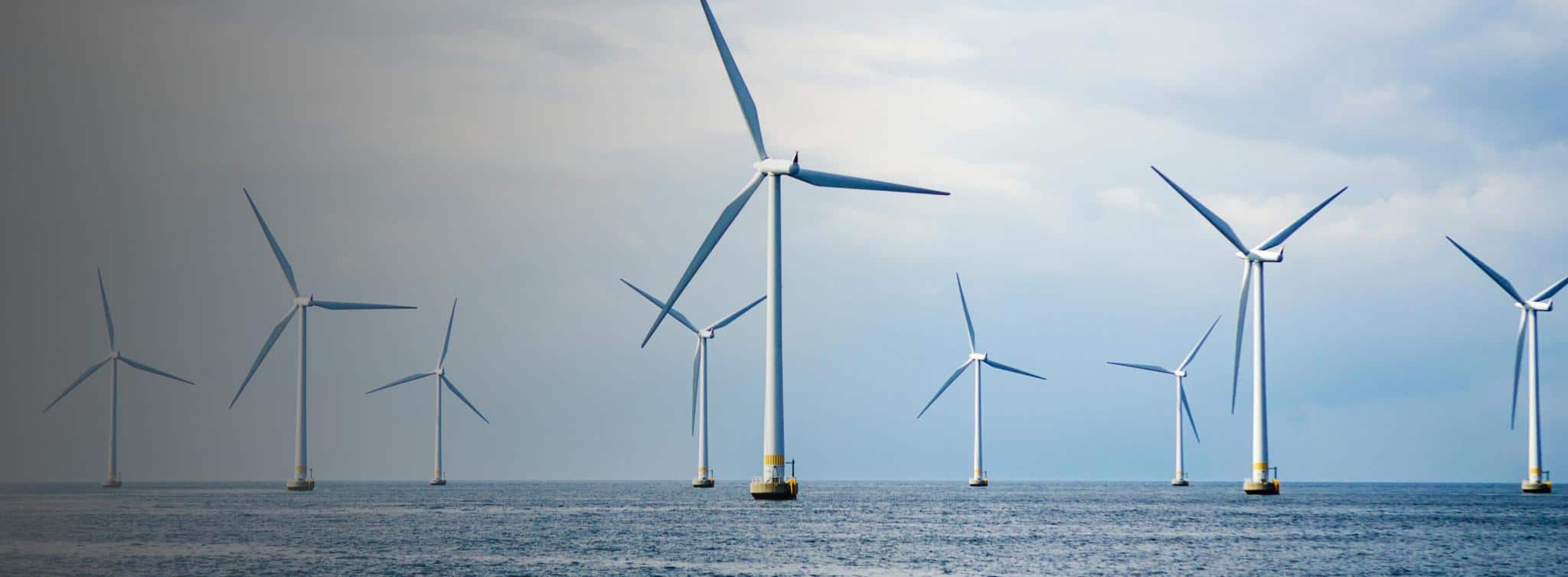 Ten large white offshore wind turbines with three blades each, standing in a blue ocean under a cloudy sky.