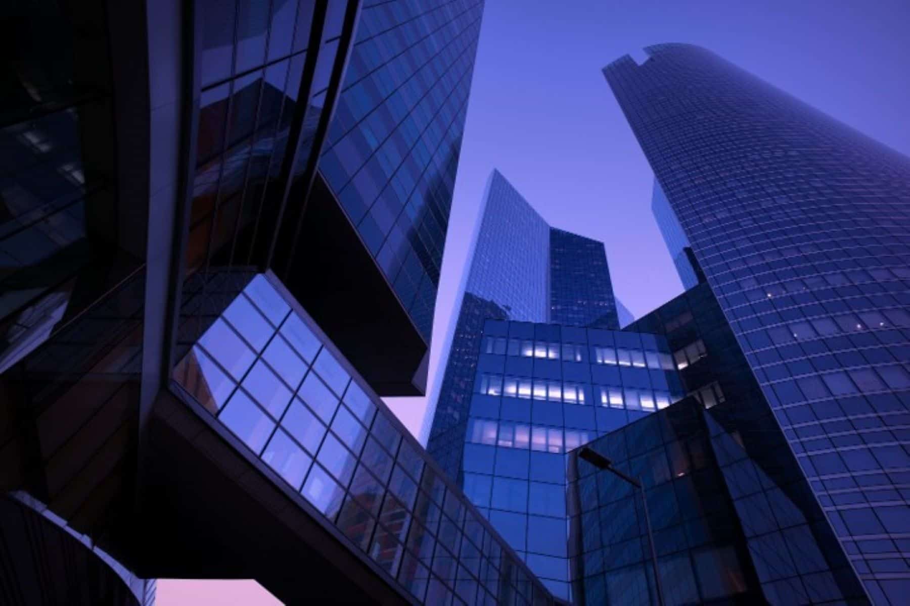 Low-angle view of modern glass and steel skyscrapers against a deep purple-blue sky, with some windows illuminated.