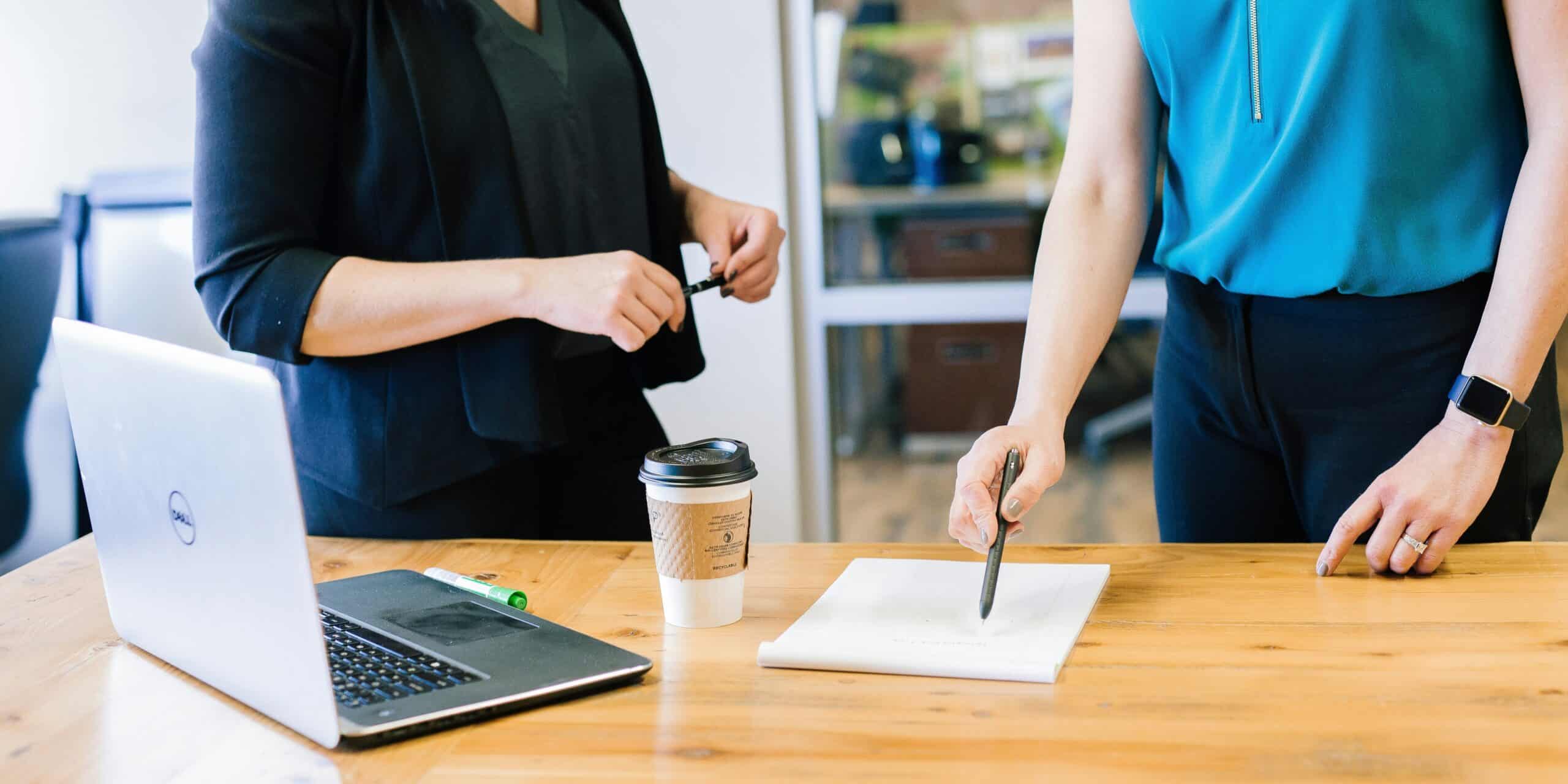 Two people discussing work at a wooden table with a laptop, coffee, and notepad.