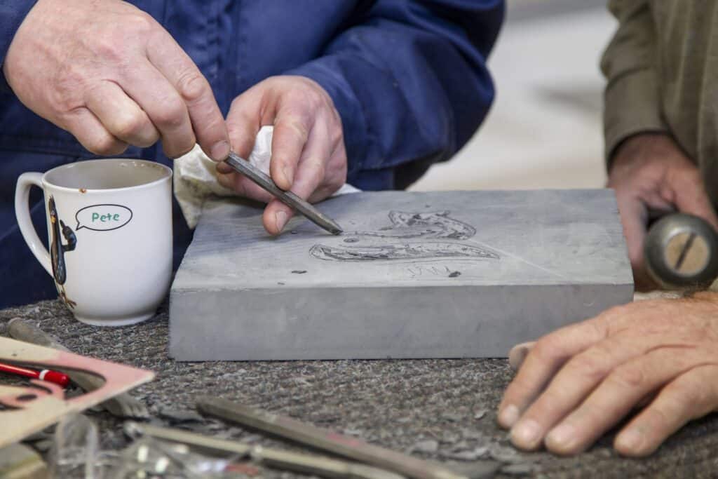 Close-up of hands carving a fish-like design into a grey stone block with a chisel, a white mug labeled 'Pete' to the left.