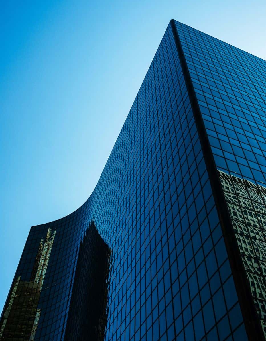 A tall, dark blue glass skyscraper with reflections, viewed from below against a clear blue sky.