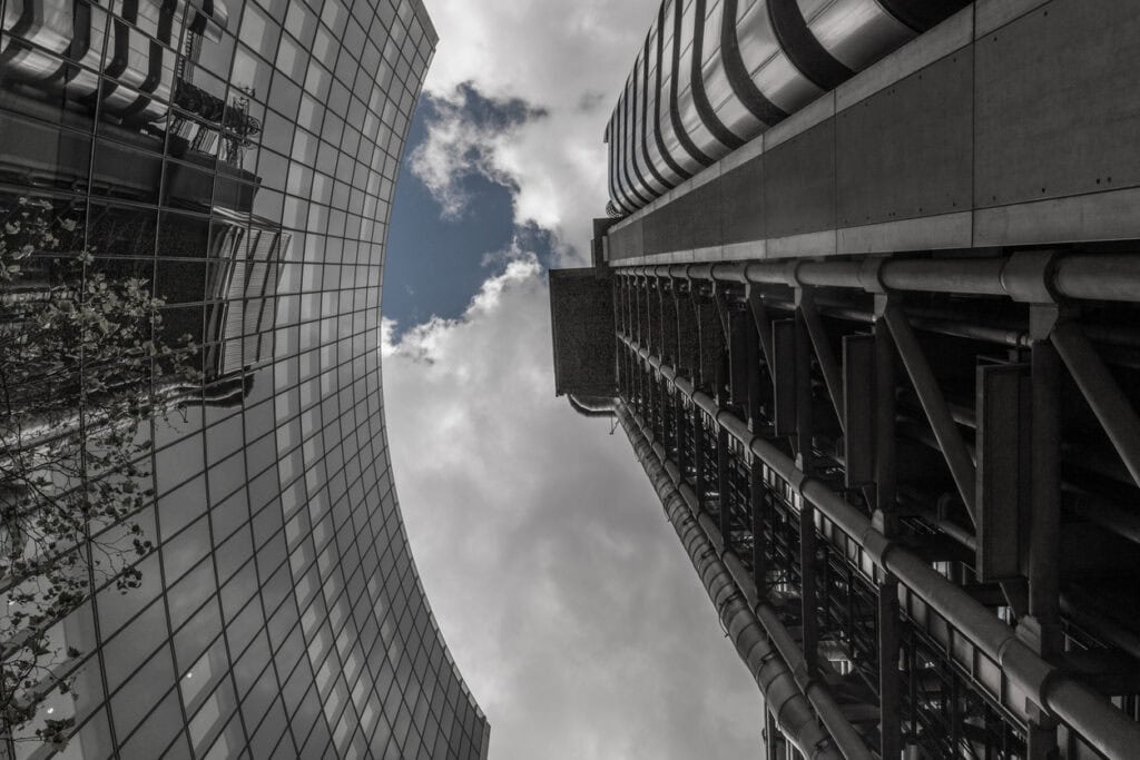 Low-angle view between two tall buildings. One has a curved glass facade reflecting trees, the other an industrial exposed structure with pipes and beams, all under a cloudy sky with patches of blue.