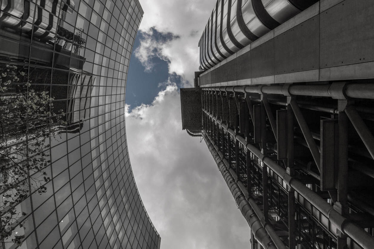 Low-angle view between two tall buildings. One has a curved glass facade reflecting trees, the other an industrial exposed structure with pipes and beams, all under a cloudy sky with patches of blue.