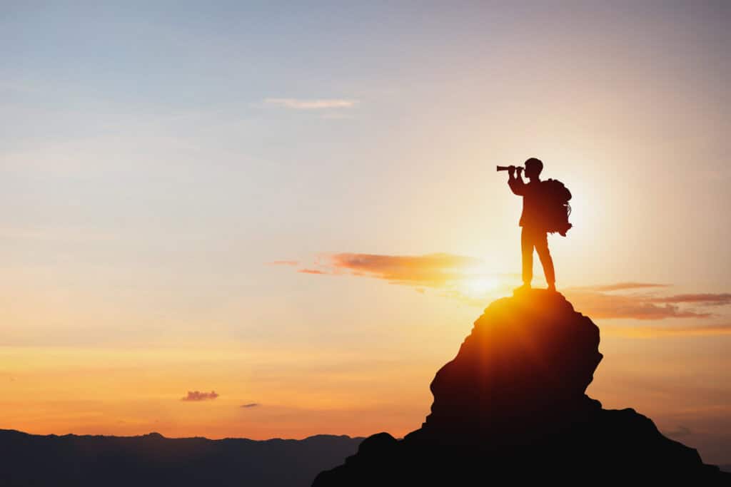 Silhouette of a person with a backpack standing on a mountain peak, looking through a monocular at sunrise or sunset.